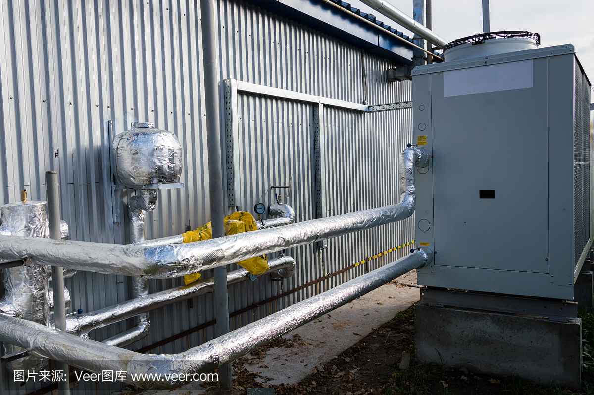在靠近現(xiàn)代制造建筑的室外地面上,與灰色工業(yè)冷卻機(jī)組相連的絕緣管道